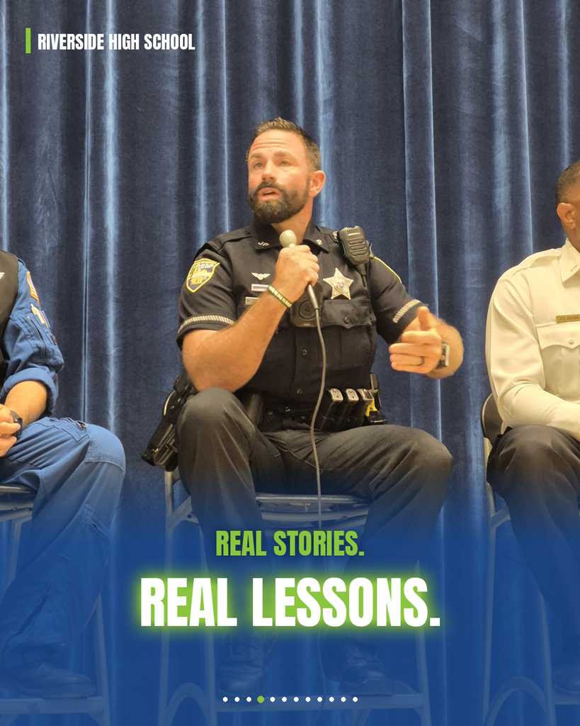 Three men in uniform seated in chairs on a stage, one holding a microphone, a blue curtain behind them.