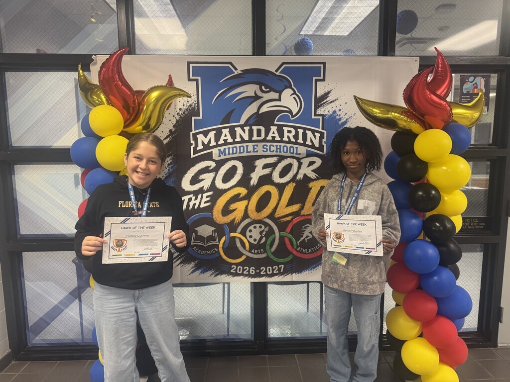Two students pose with certificates in front of a banner and balloons in front of a window.