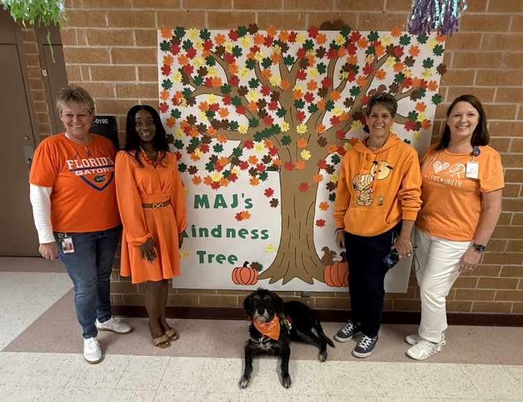 Four adults in orange shirts stand in a hallway with a black dog, near a brick wall with a poster of an autumn tree.