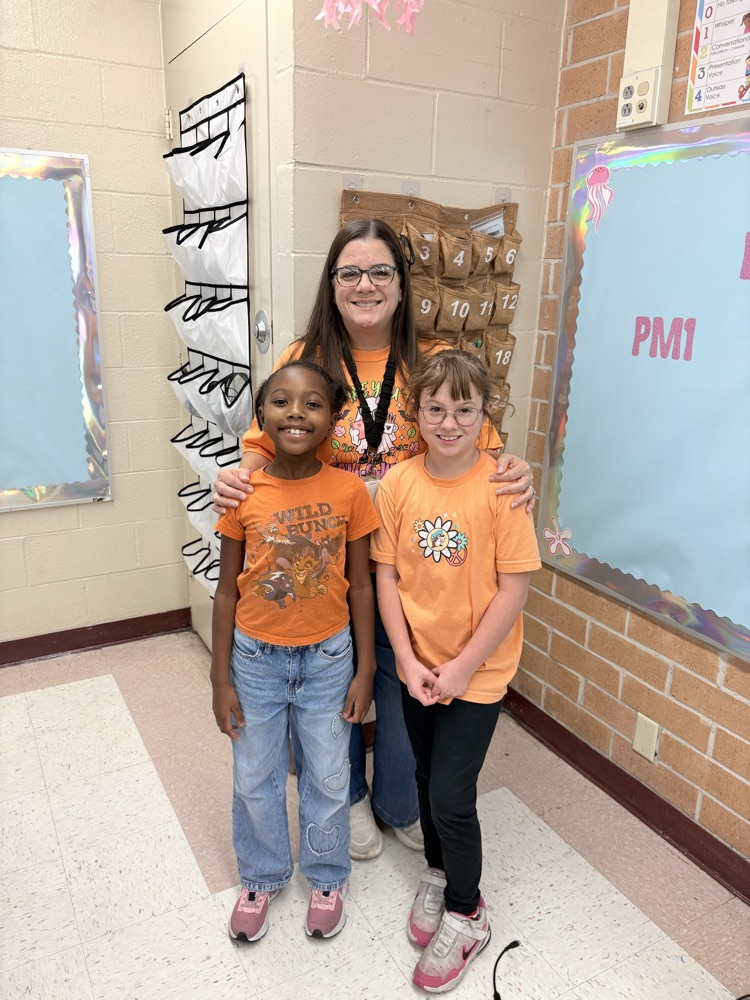 Three children, two in orange shirts, one with glasses, standing in front of brick wall and bulletin boards.