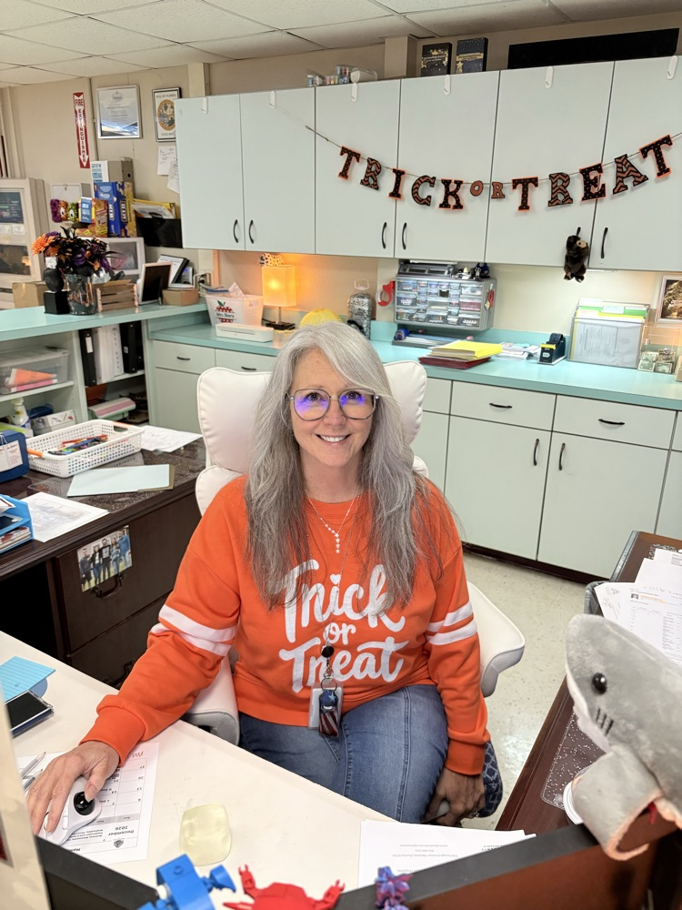 Woman in orange sweatshirt sits at a desk with papers and a stuffed shark.
