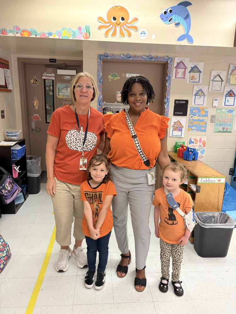 Two adults and two children stand in a classroom hallway. The adults wear orange shirts, while the children wear orange t-shirts.