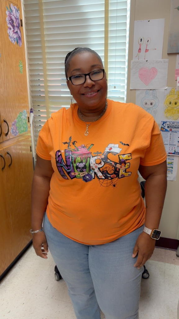 Person wearing orange shirt with Nurse print, glasses, necklace, and bracelet, standing by a window.
