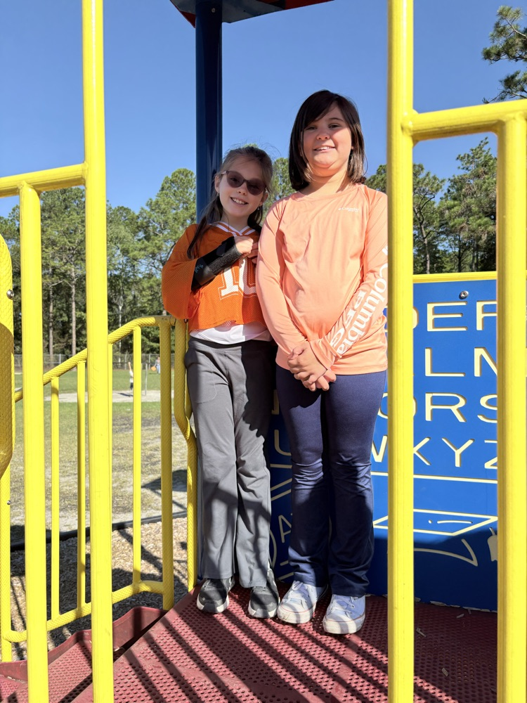 Two girls in orange shirts pose on a playground with yellow railings and letters.