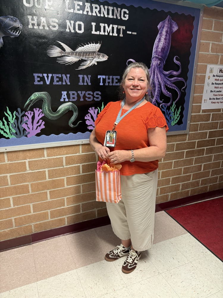 Adult wearing orange shirt and khaki pants standing next to a brick wall with fish mural and red rug.