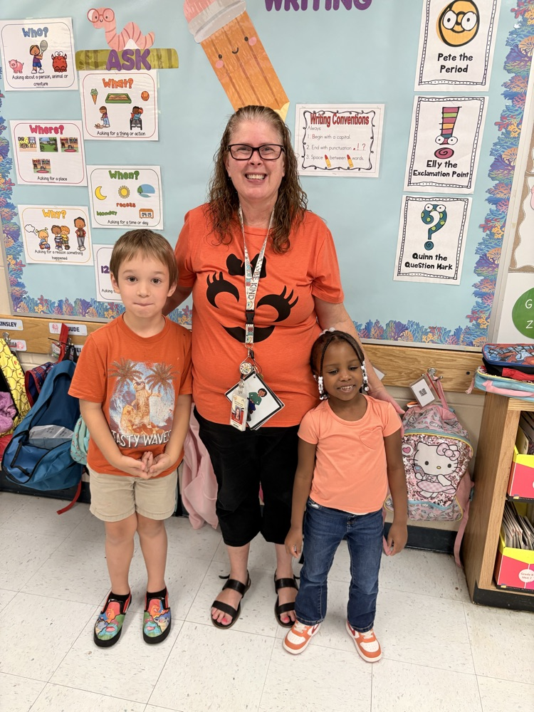 Woman and two children in front of a bulletin board with colorful papers. The woman wears glasses and an orange shirt.