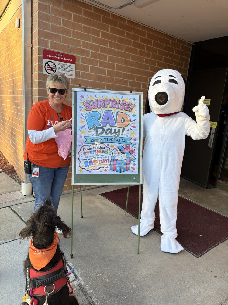 Person in orange shirt holding sign next to mascot in white costume standing by sign, dog sitting.
