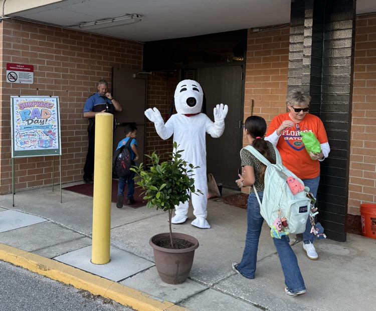 Adults and children, including one in a dog costume, walk past a potted plant. A police officer stands in the doorway.