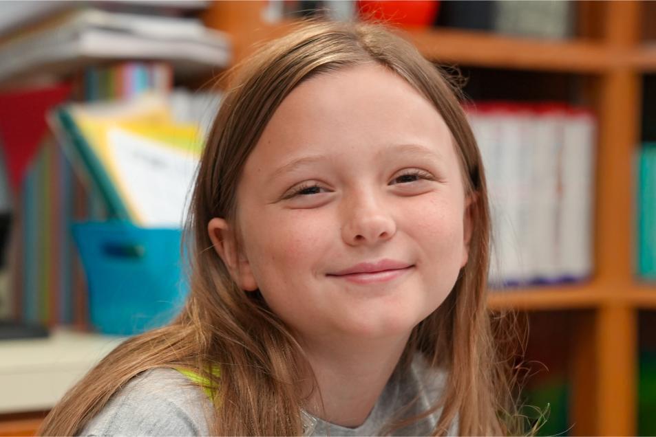 A young girl smiles in a classroom, with a bookshelf behind her filled with books and a blue bin.