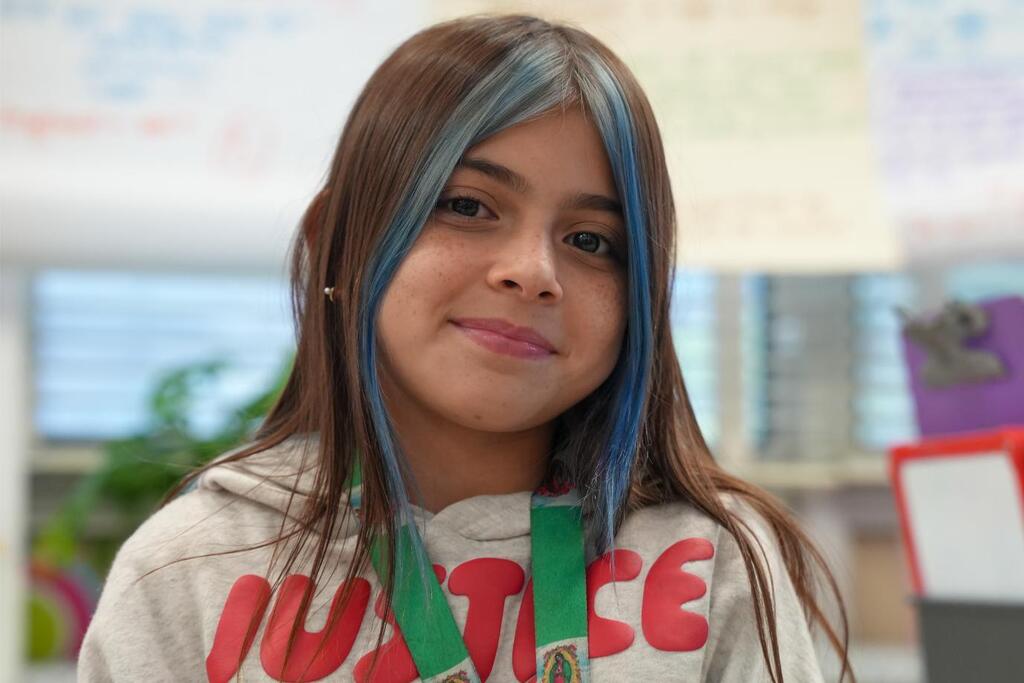 A girl in a gray hoodie, smiling, with blue hair, wearing a green medal around her neck.