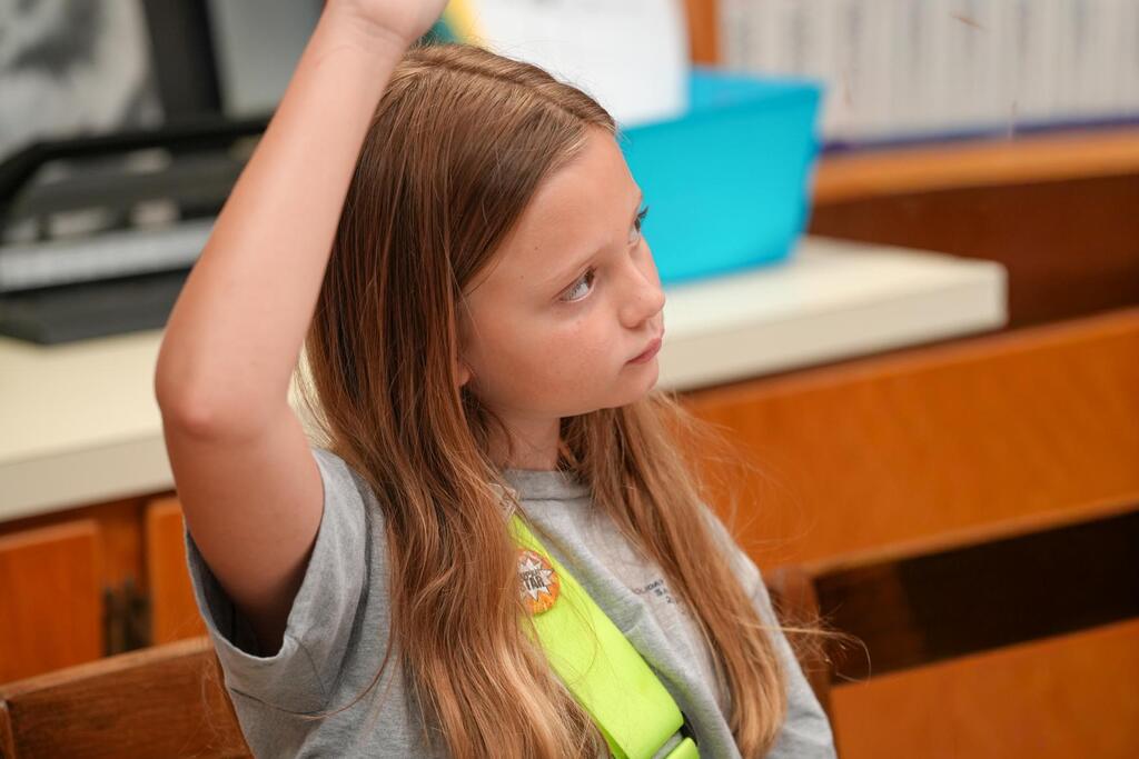 A girl with long hair raising her hand in a classroom with a blue bin on a table.