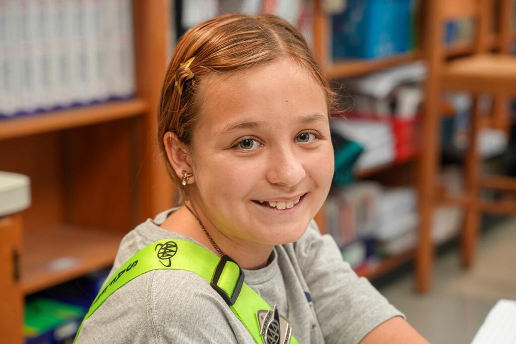 A girl sits at a table, smiling, with a yellow safety harness. Behind her is a bookshelf filled with books.