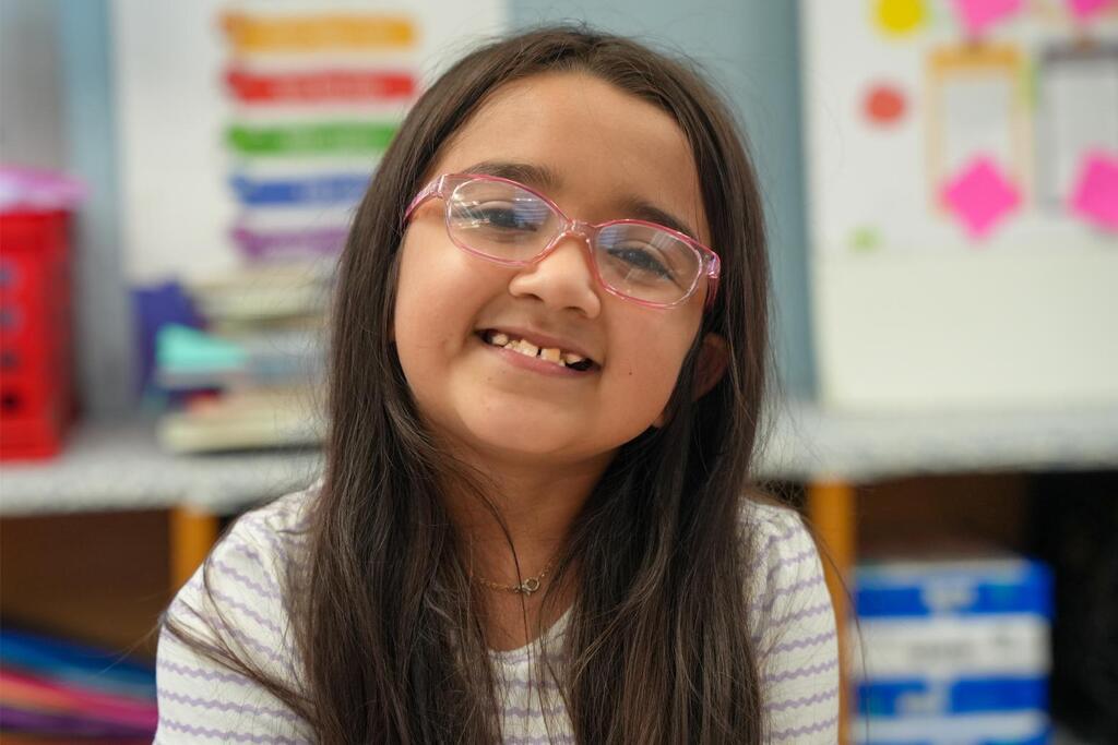 Girl with glasses smiles at camera in classroom; desks, books, and a whiteboard with colorful dots.