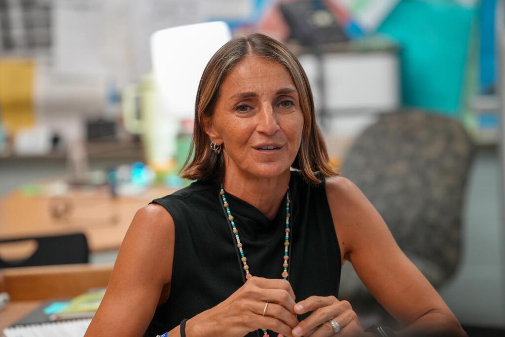 Woman in black sleeveless top sits at desk with papers, gesturing with hands while facing forward.