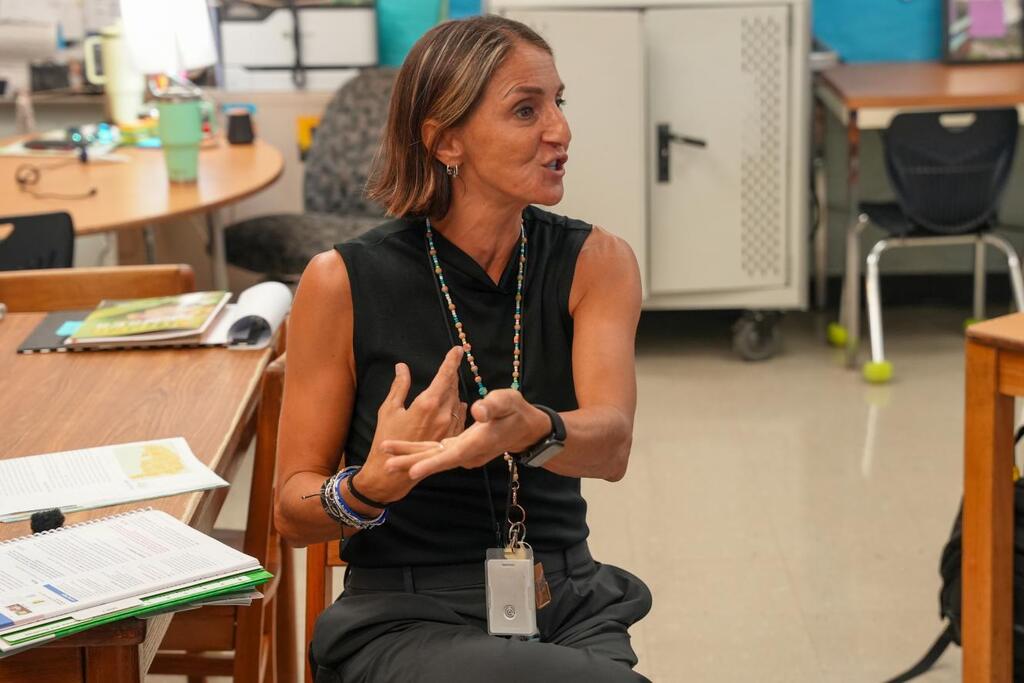 Woman seated in classroom, gesturing with hands while speaking. Papers, notebooks, and a backpack are on the desk.