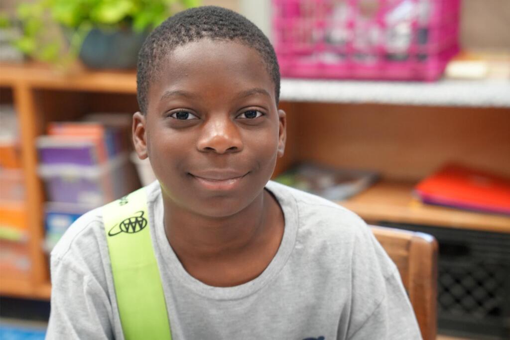 A smiling boy wearing a gray shirt and a green sash sits in a chair against a bookshelf background.