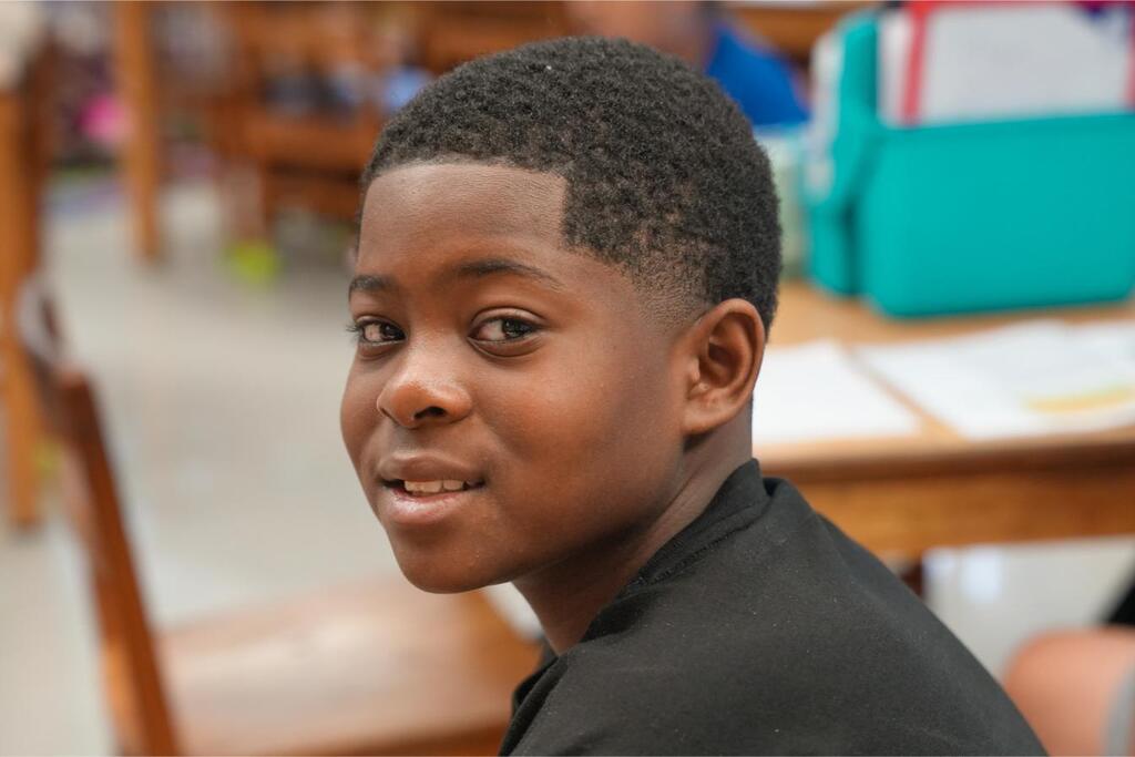 Child with short hair, smiling, sitting in classroom. Desk, chair, and bag in background.