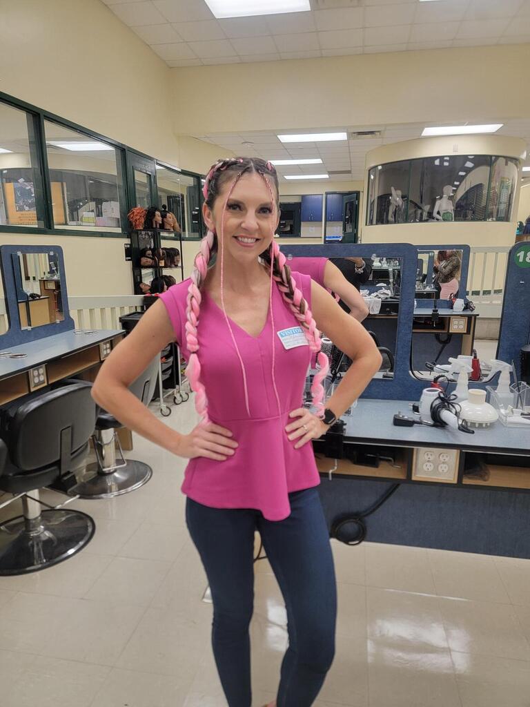 Woman in pink shirt and braids stands in room with salon chairs and tables in background.