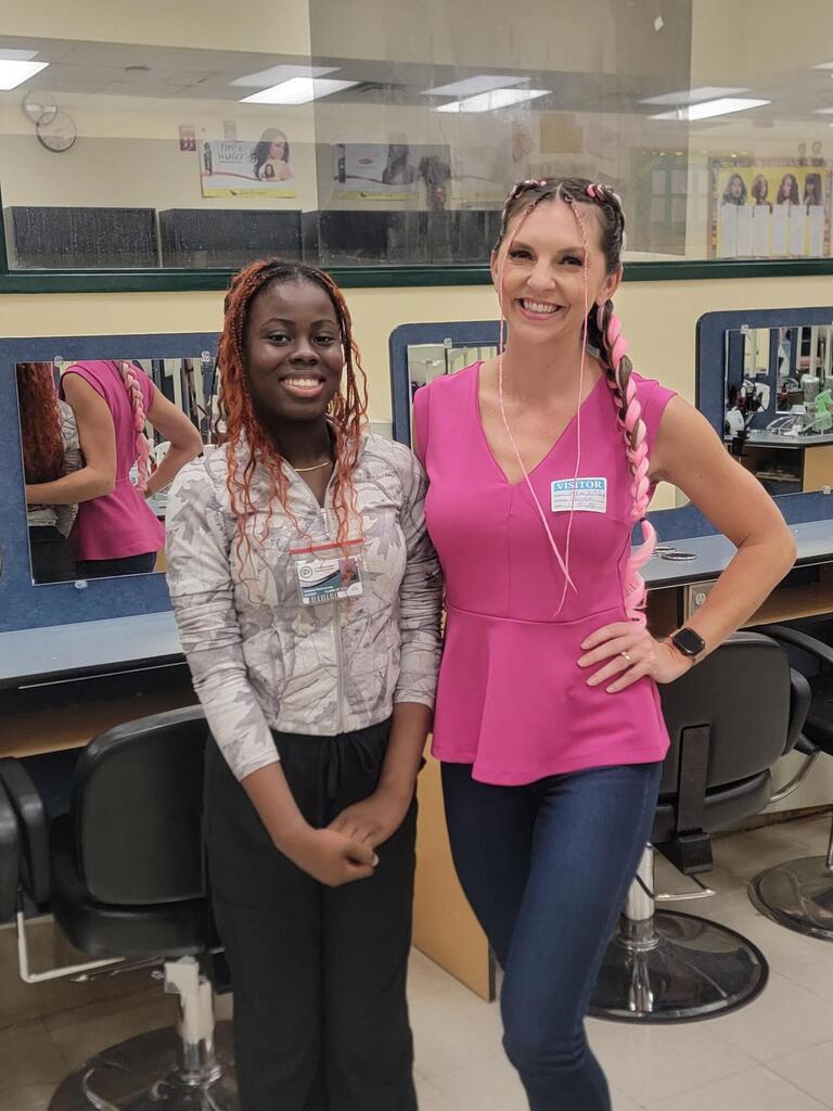 Two women standing in a salon; one with curly hair, wearing glasses, standing next to a smiling woman with braided hair.