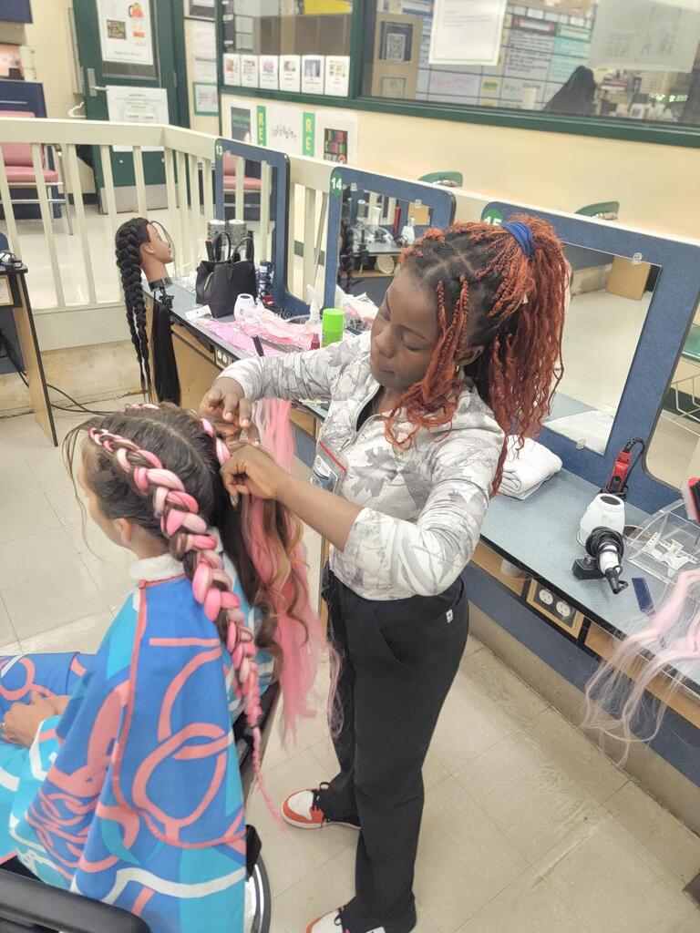 A student braids another student's hair in a salon chair while seated at a counter.