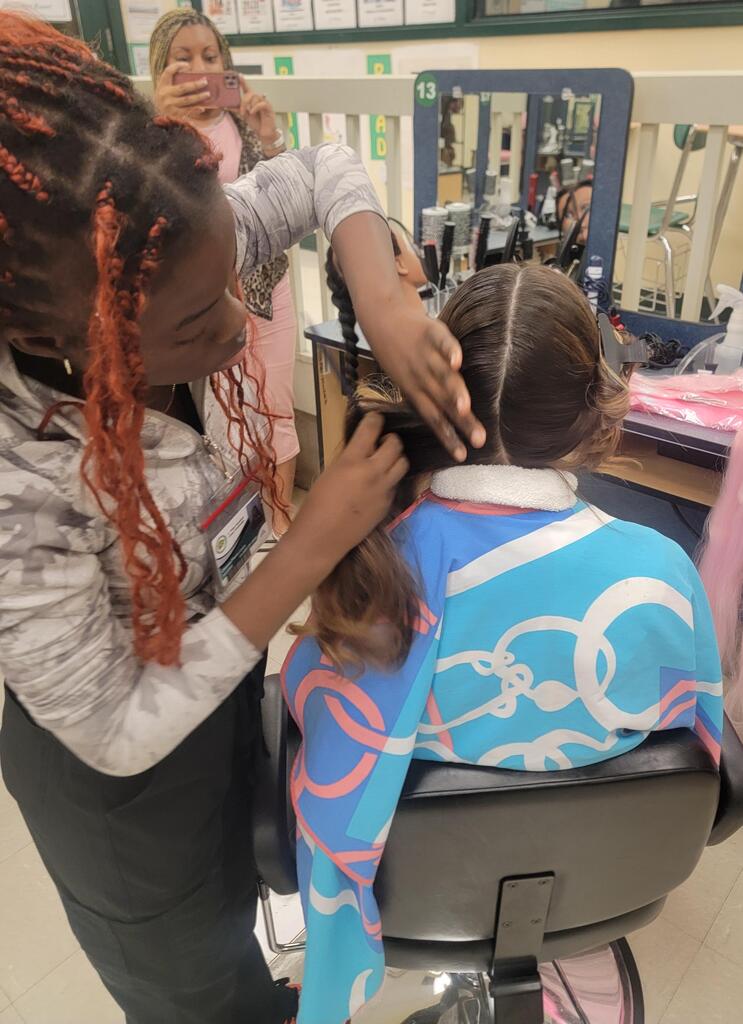A girl with braided hair styles another girl's hair in a chair in a room with mirrors.