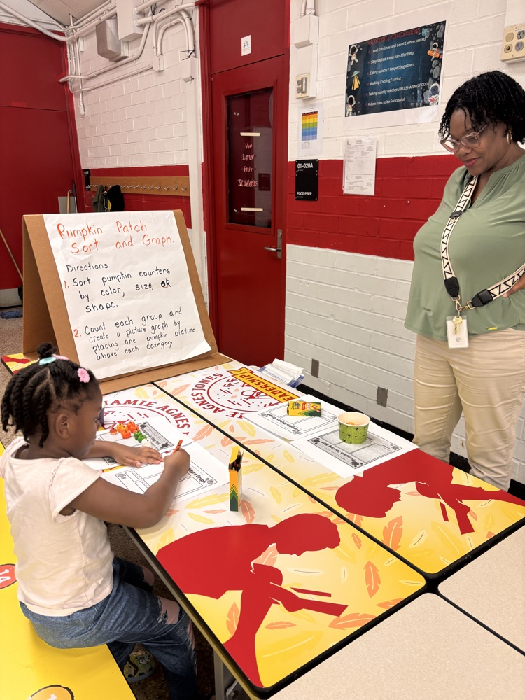 Girl seated at a table with a drawing on it. Woman stands behind her, looking at her work.