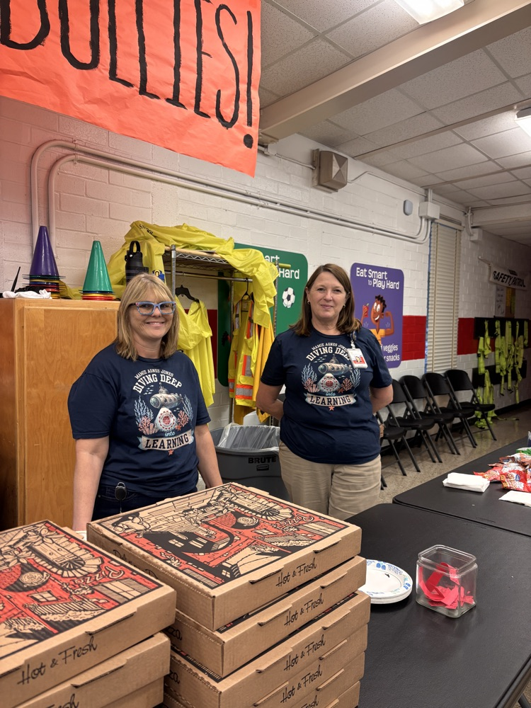 Two women stand behind a table with pizza boxes, a glass jar, and a plate.