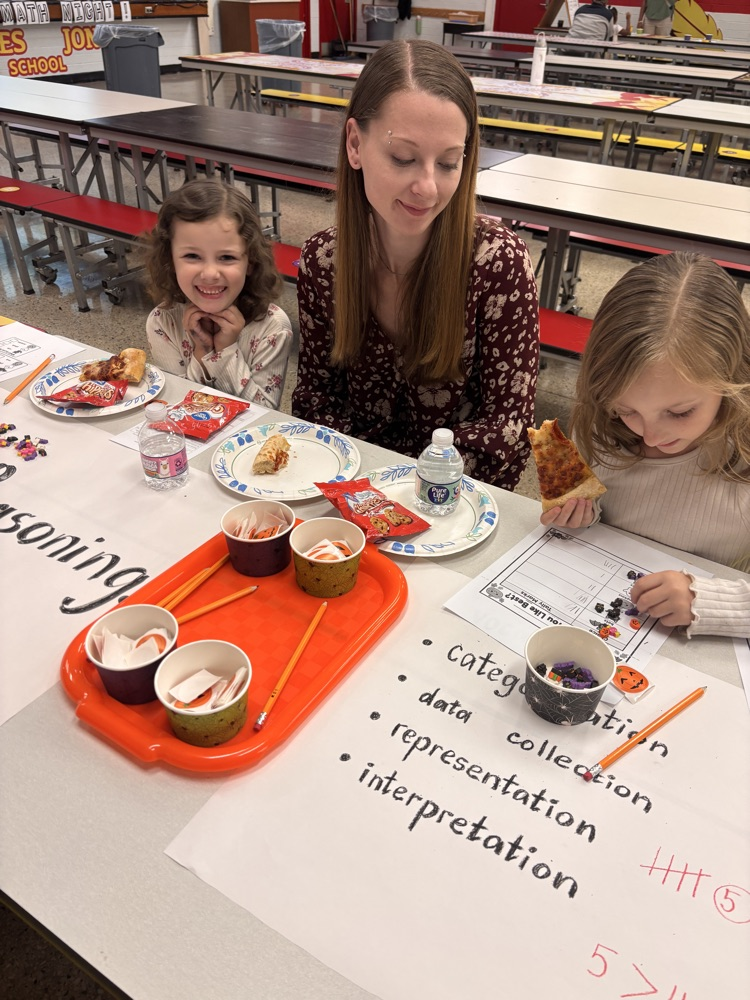 A woman and two girls eat food while sitting at a table with papers and writing tools.