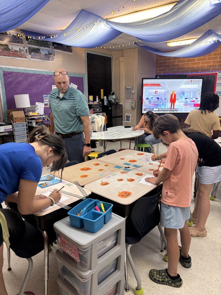 People in a classroom; two adults and four children working on a table, with a teacher and a screen