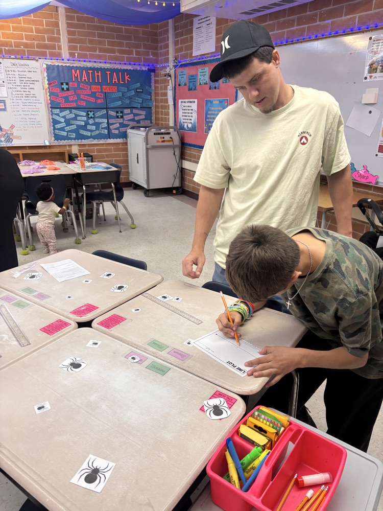 Boy using colored pencils at desk, writing with another boy in cap nearby. Walls have bulletin boards and signs.
