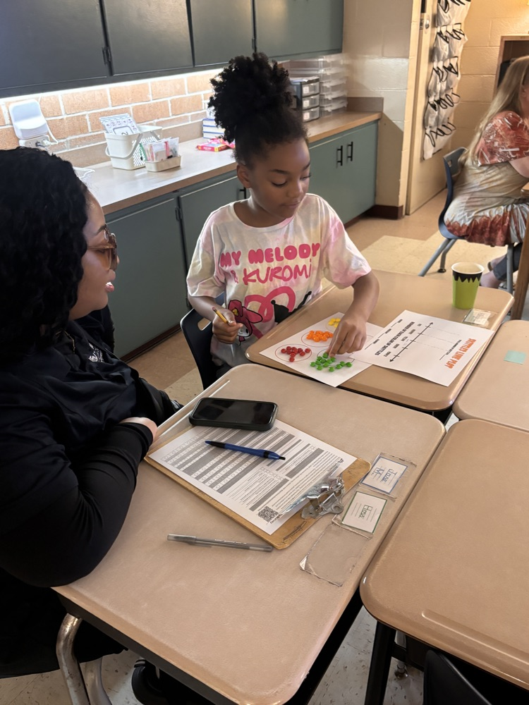 Woman sitting at a desk helps a child with a worksheet. The child holds a paper with a red cross.