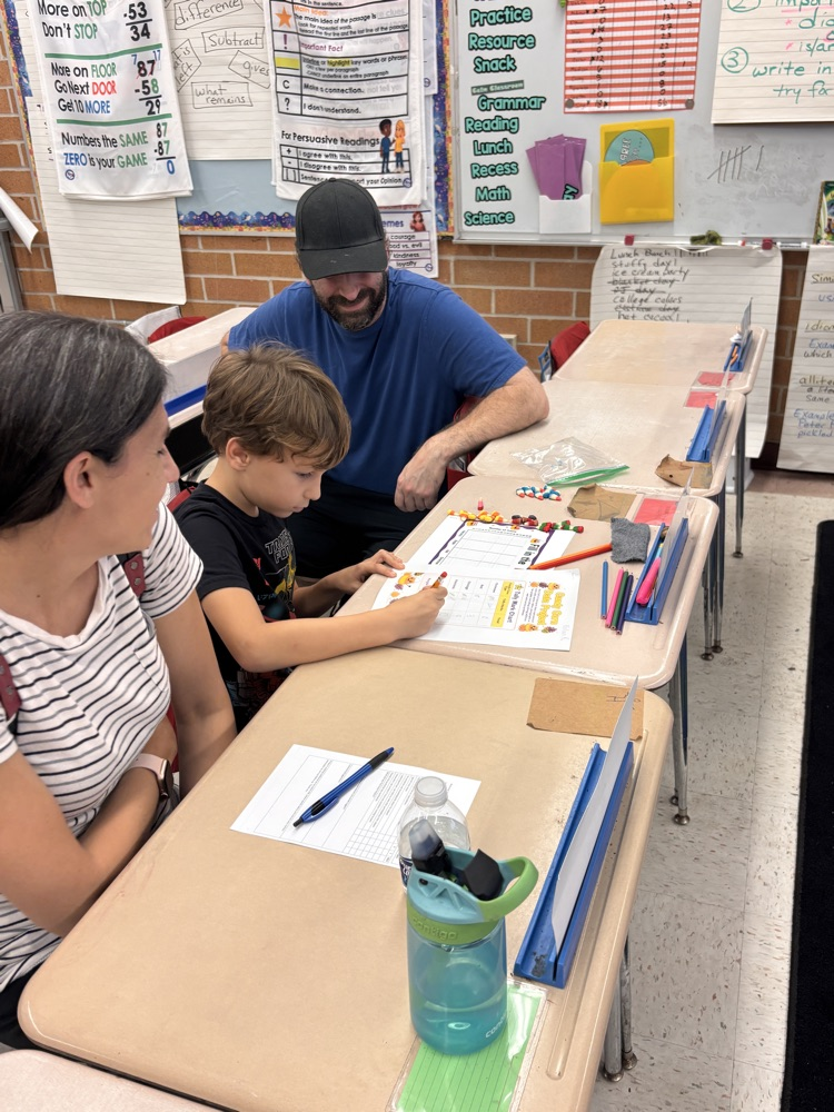 Teacher assists a student with coloring a worksheet. Woman observes from behind. Supplies on table include pencils, markers, pens, and paper.