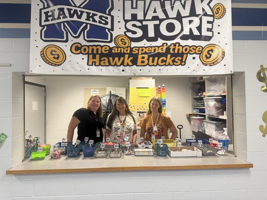 Three women smiling behind the counter of a school store. Above them hangs a large white banner that reads Hawks Hawk Store - Come and spend those Hawk Bucks! with illustrations of gold coins with dollar signs. On the counter in front of them are many small baskets and bins filled with colorful small prizes and school supplies, each labeled with prices like $1, $2, and 2 for $1. The store is set in a small room with white brick walls and shelves with supplies in the background.