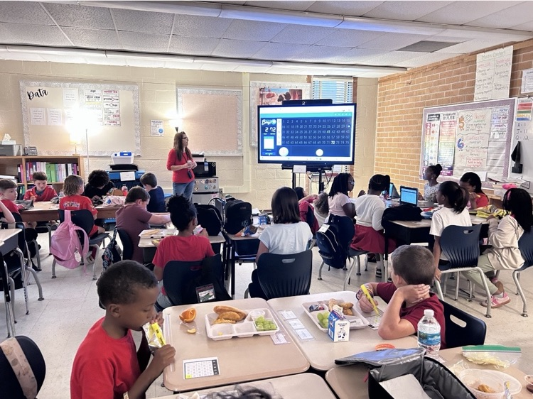 Children sit at desks in a classroom, eating. A teacher stands by a screen, and a whiteboard is behind her.