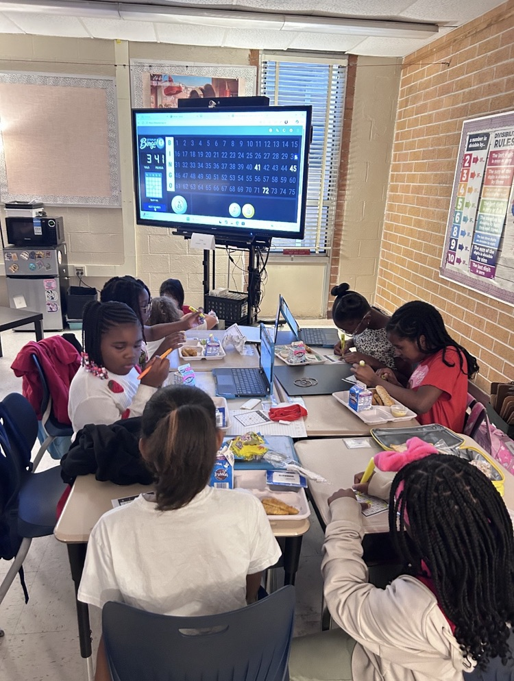 Students seated at tables in a classroom, some using laptops, one with a tablet, and food items on the tables.