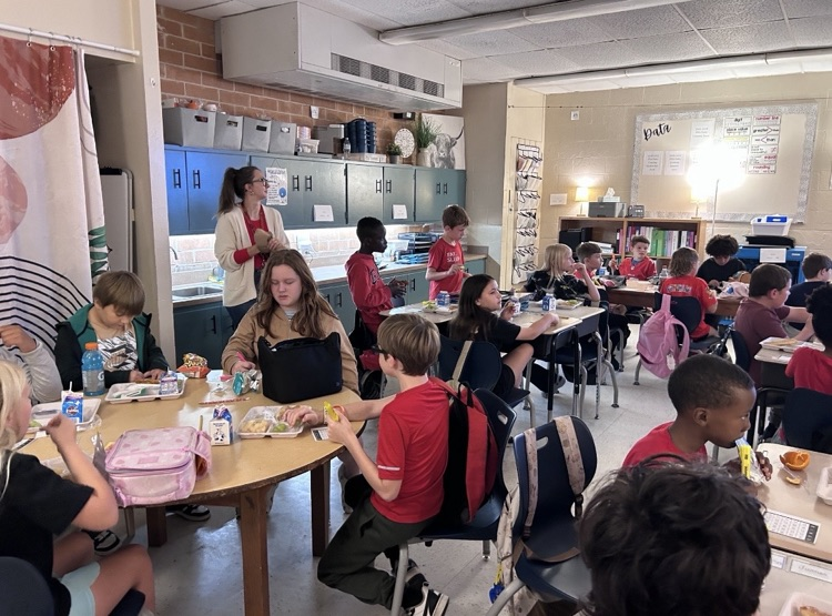 Children seated at tables in a classroom with adults nearby. A woman with long hair stands behind a table.
