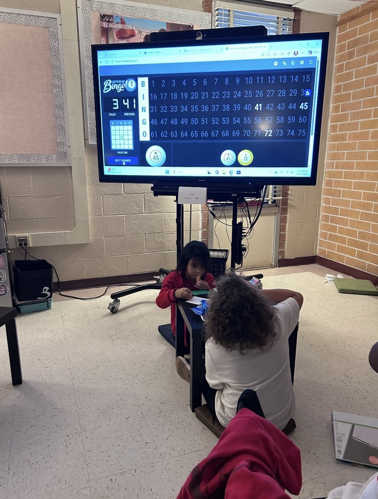 Two students, one with a curly brown head of hair, are seated at a table playing a game on a monitor.