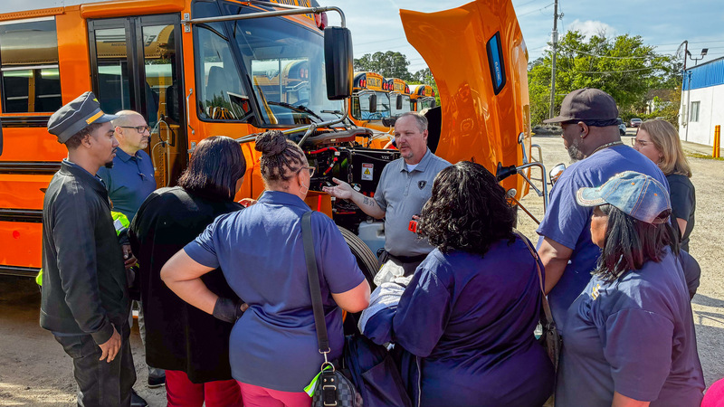 School bus personnel stand outside in front of an Electric Bus as a trainer does a demonstration