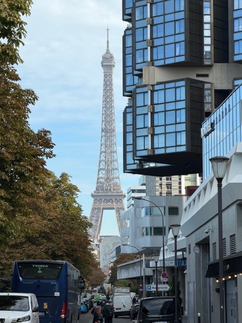 Modern buildings with glass facades. The Eiffel Tower stands tall in the background. A road with cars, buses, and motorcycles.