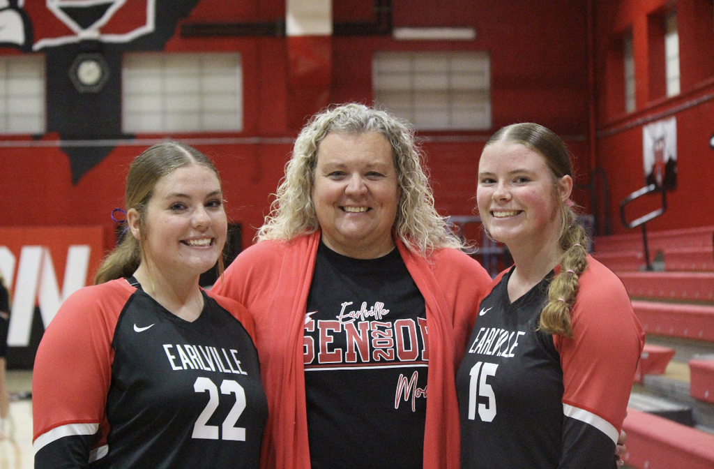 Three women in sports uniforms posing in front of red gym bleachers, one with a pink jacket.