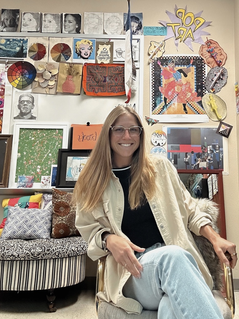Woman with blonde hair and glasses sits in a chair in a room filled with artwork.