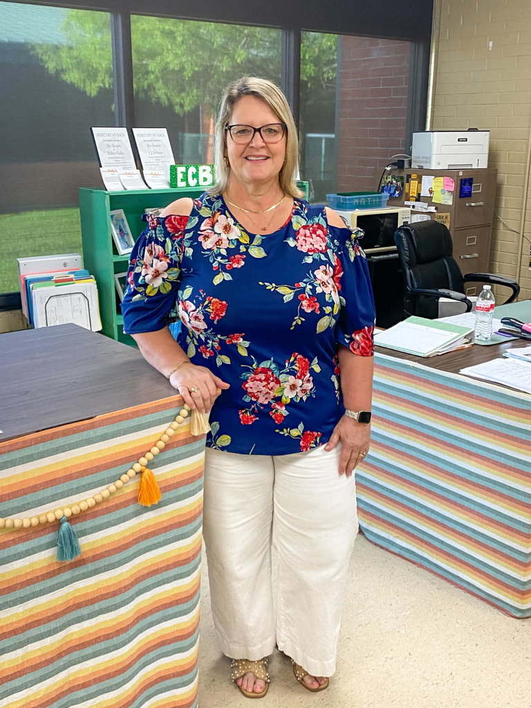 A woman stands in an office with a colorful striped desk behind her. She wears a blue floral blouse and white pants.