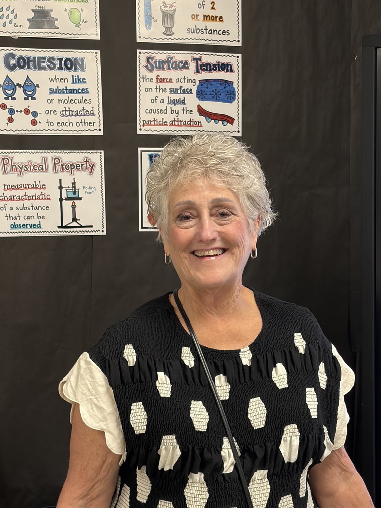 A woman with short hair smiles in front of a wall with educational posters.