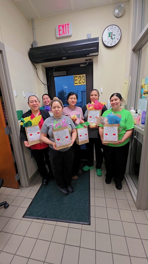 Six women stand in a corridor, each holding a gift bag with a red heart. A green rug and a clock are visible in the background.
