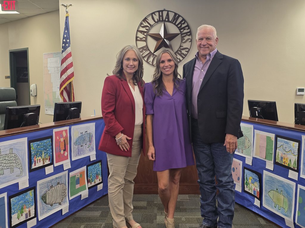 Three people stand near a wall of artwork in an office. A woman in red stands between two men.
