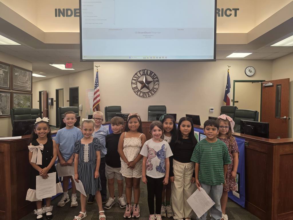 A group of children in a room with a presentation screen above, holding certificates. A Texas flag is displayed.