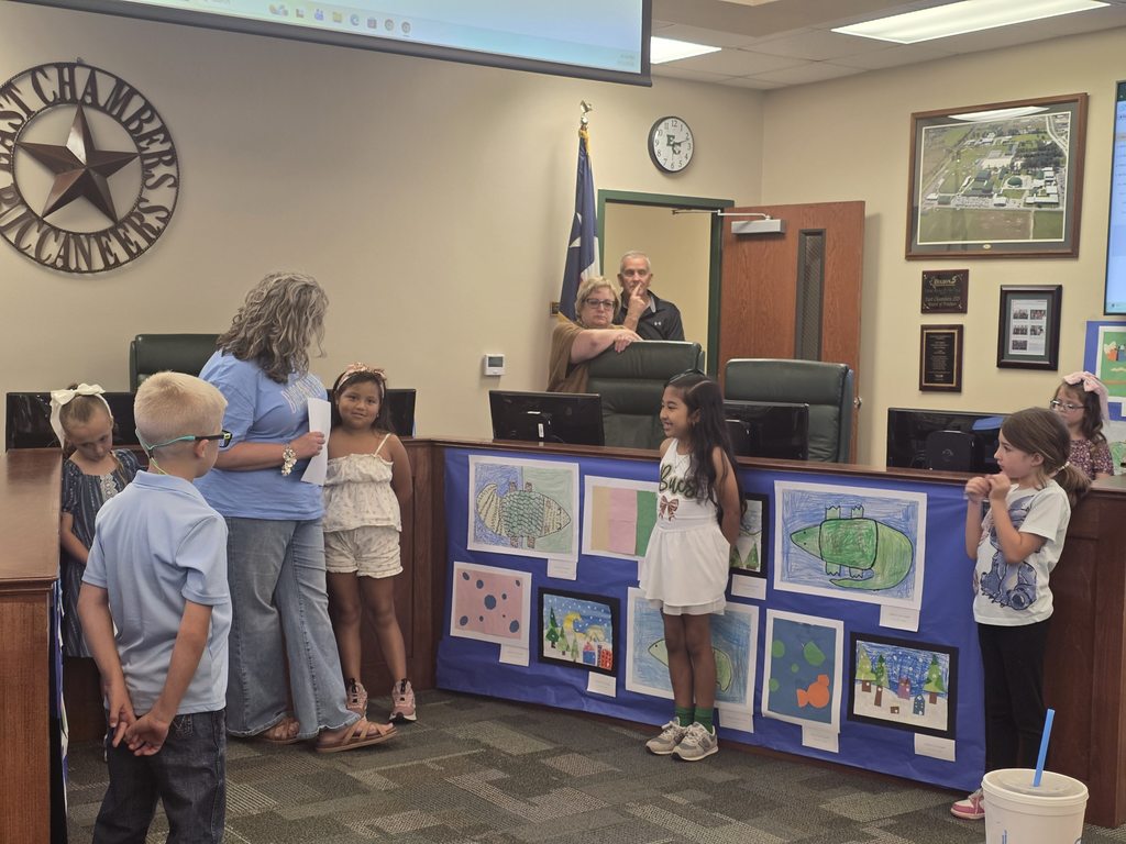 Children and adults in a room with a backdrop of artwork. A person stands behind a desk.