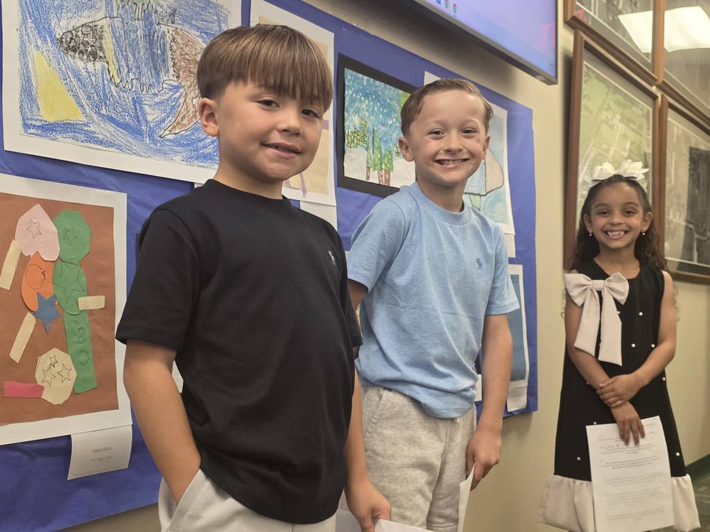 Three children stand near art pieces on a wall. A boy in black, a boy in blue, and a girl in white.
