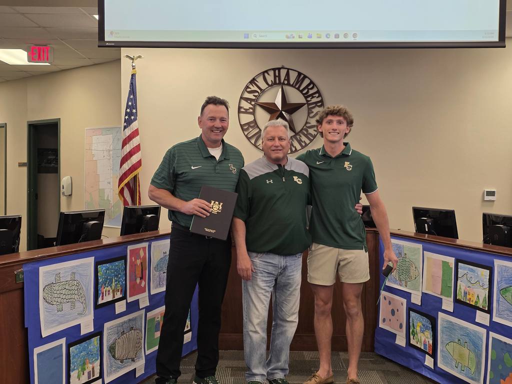 Three men in green shirts stand before a desk with artwork in a room with a flag.