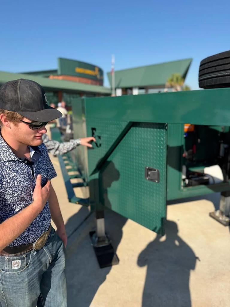 Person in a hat and sunglasses standing by a green machine, pointing to a door on it.